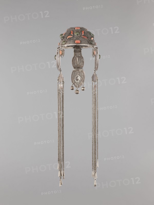 Headgear ornament coif, 19th century, Mongolia. Women's headgear / head ornament made of silver with ear cuffs; filigree. Height 57.0 cm, diameter 14.0 cm., An ornate metallic headdress with a domed top decorated with oval and rounded cabochon stones in shades of coral and green, featuring intricate filigree and textured silverwork; two curved side pieces extend downward from the dome and connect to a central vertical pendant composed of linked engraved segments and small spherical bells, with long delicate silver chains hanging from each side ending in small cylindrical metal cones; the colors visible are silver, coral, green, and subtle variations of gray against a neutral light-gray background.