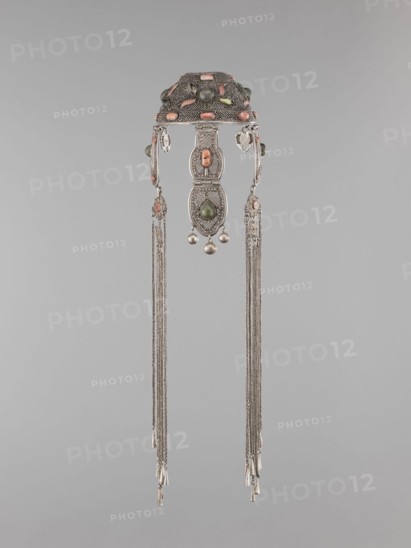 Headgear ornament coif, coif, jewelry, Mongolian art, artistic crafts, headgear, head ornaments, women's ornaments. Made of silver with ear cuffs; filigree. 19th century, Mongolia. Clothing accessory. Height 57.0 cm, diameter 14.0 cm., An ornate metallic headdress piece hangs against a plain light gray background, featuring an arched top section decorated with rounded colored stones in shades of coral and green set into textured silver metal; dangling from the top are three linked filigree panels with an oval green stone in the central lower panel and small spherical silver beads suspended beneath it; two long groups of fine silver chains extend downward from either side, each ending in small tapered metallic tassels, and there are small coral-colored accents and green beads visible on the side elements, with overall colors including silver, coral, green, and the neutral gray of the background.