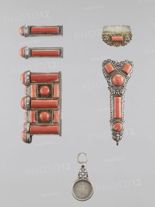 Braid ornament, braid ornament jewelry, artistic craft; braid ornament for women's dress, 19th–20th century, Mongolia; materials: jewelry; height 3.0 cm, width 7.8 cm., A vertical arrangement of several ornamental metal pieces set against a pale gray background, featuring a group of jewelry-like objects with coral-colored stones framed in silver-toned metal, including a rectangular cluster with rounded coral cabochons and long coral bars, a trio of slim rectangular pieces each capped with a rounded coral end, an ornate pendant-like piece with a heart-shaped top and vertical coral panels, a small scalloped metal element with a coral rectangle, and a round coin-like medallion suspended from a decorative silver loop; visible colors include coral, silver, and pale gray.