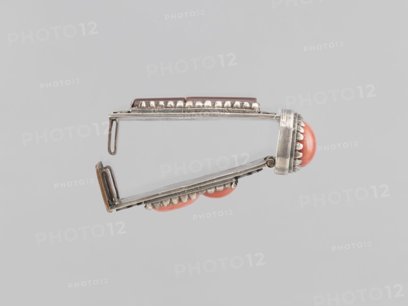 Braid clasp, braid ornaments, clasps, jewelry, Mongolian art, artistic crafts, women's ornaments, late 19th century, Mongolia, jewelry; height 2.9 cm, width 2.3 cm, length 7.4 cm, A metallic hinged object shown against a light gray background, photographed from the side, with a silver tone and slightly tarnished finish; the object has decorative scalloped metal edging and a few rounded coral-colored cabochon elements set into the metal; one coral element is prominent at the hinged end and a couple more are visible along the lower edge; the hinge and clasp components are visible with thin silver bars and a small looped hook; overall colors visible include silver, coral, and light gray.