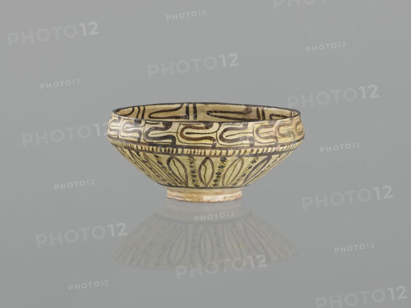 Bowl, 10th century, Nishapur, Iran; Persian Islamic ceramic vessel decorated with images of birds and palmettes; weight 1.11 kg, height 12.1 cm, diameter 26.0 cm., A shallow bowl with a rounded body and small foot, decorated all around with curving and leaflike painted motifs; the bowl shows yellow, black, brown, beige, and a hint of reddish tones, and it sits on a smooth gray surface that produces a soft reflection of the bowl.