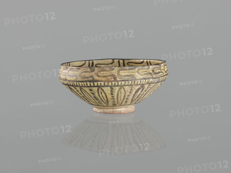 Bowl, 10th century, Nishapur, Iran; Persian Islamic ceramic vessel decorated with images of birds and palmettes; weight 1.11 kg, height 12.1 cm, diameter 26.0 cm., A shallow bowl with a rounded rim and a small foot, decorated with painted geometric and leaf-like patterns, shown against a plain gray background with a soft reflection below; visible colors include beige, black, brown, cream, and hints of ochre.