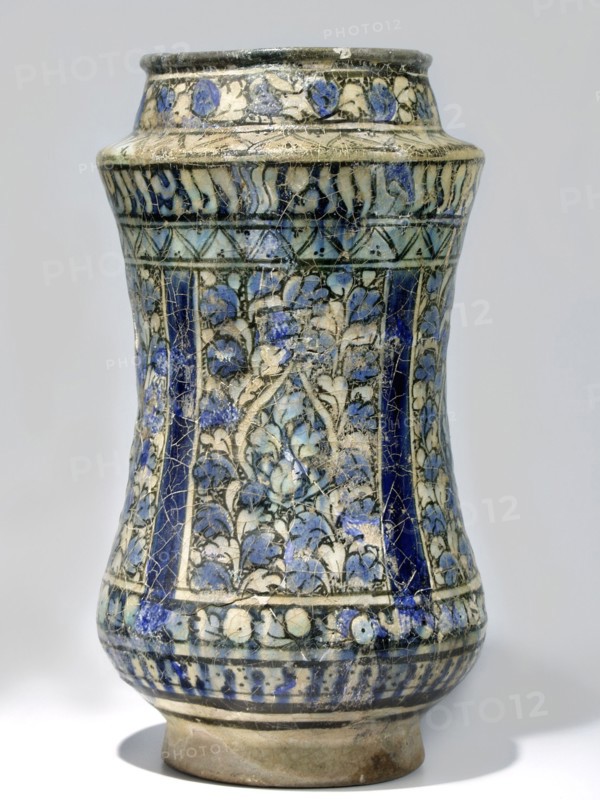 Albarello, 14th century Iran, Persian, Islamic art. Ceramic albarello vessel with floral/plant motifs, weight 0.99 kg, height 25 cm, diameter 13.5 cm., A tall ceramic vessel with a slightly flared rim, a bulging midsection, and a narrower base, decorated all over with an intricate painted pattern of floral and vine motifs; the surface shows a network of fine cracks. The visible colors include shades of blue ranging from deep cobalt to lighter sky blue, off-white or cream, black outlining, and areas of beige or light brown on the unglazed base. Several vertical panels of dense floral decoration alternate with darker blue vertical bands, and there is a band of repeating floral motifs near the top and another decorative band near the bottom. The vessel sits on a short, unglazed foot and casts a soft shadow on the surface beneath it.