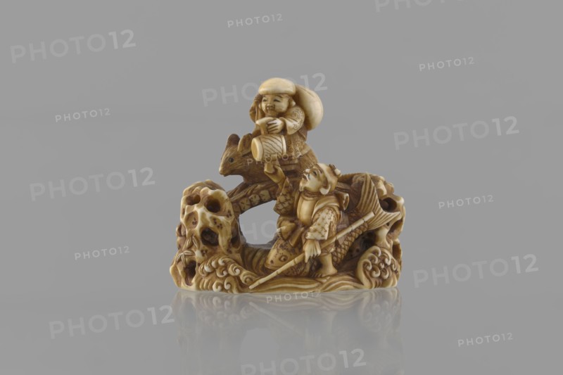 Netsuke in the shape of Daikoku and Ebisu, Ryôji fl. ca. 1886–1900? sculptor; author unknown. Japanese, Meiji period 1868–1912. Carved miniature netsuke katabori depicting Daikoku and Ebisu from the Seven Gods of Good Fortune: Daikoku sits on a rat holding a hammer in his left hand with a bag on his back; Ebisu holds a fishing rod and rides a giant fish. Finely carved in a miniature rocky landscape with a bridge, showing detailed facial features, fish scales, bamboo leaves on openwork rocks, rat fur, and rat’s eyes set with microscopic black-dyed bone inlays. 4th quarter of the 19th century, Japan. Height 3.9 cm; width 4.0 cm., A small carved ivory-colored figurine depicts a scene with a few humanlike figures and an animal on an ornately textured base; the carving shows a standing figure holding a long pole and wearing patterned clothing, a second seated figure above holding a rounded object, and an animal with pointed ears nestled between them, all carved with detailed patterns, scale...