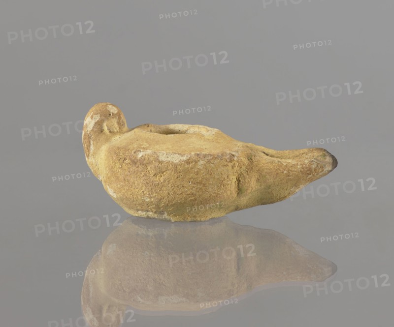 Oil lamp, Islamic art, Islamic and Arabic culture, utility ceramics, Arabic ceramics, everyday life in the Middle East, Egyptian ceramics, 8th–9th century, Egypt place of origin, oil lamp, height 9.4 cm, width 5.7 cm, depth 4.1 cm, A weathered oil lamp shaped like a shallow bowl with a pronounced spout and a rounded handle, shown on a reflective surface that creates a soft mirror image; the lamp has a rough, uneven texture with patches of lighter wear and small areas of greenish discoloration, and the visible colors include shades of tan, ochre, brown, pale yellow, and hints of green against a neutral gray background.