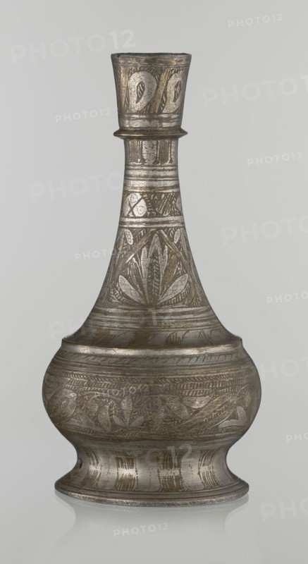 Bottle, Islamic art, Arabic culture, geometric and plant motifs, neck slender toward the top ending with a straight spout. Engraved patterns painted with golden-brown paint. Early 20th century, Middle East, vessel, height 21.6 cm, width 10.5 cm., An ornate metal vessel with a narrow neck and flared rim sits upright against a plain light gray background; the vessel displays an overall metallic silver-gray color with areas of darker gray and subtle brownish or bronze tones, and its surface is covered in engraved decorative patterns including leaf-like and curved motifs arranged in bands around the neck, shoulder, and bulbous body, with linear ridges and horizontal bands separating the patterned sections; the base flares outward and shows vertical engraved lines and a slightly reflective surface that creates a faint mirrored reflection on the surface beneath.