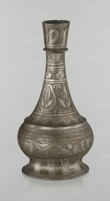 Bottle, Islamic art, Arabic culture, geometric ornament, plant motifs; neck slender toward the top ending with a straight spout; engraved patterns painted with golden-brown paint; early 20th century, Middle East; vessel; height 21.6 cm, width 10.5 cm, The image shows a single metal vessel with a bulbous base, a narrowing neck, and a flared rim, standing against a plain light gray background. The vessel has engraved and incised decorative patterns covering most surfaces, including repeating leaf-like and geometric motifs. The metal surface appears aged with a patina, showing shades of silver-gray and darker gray, along with areas of brownish or bronze discoloration. The reflections and subtle highlights emphasize the metallic texture and the engraved details.