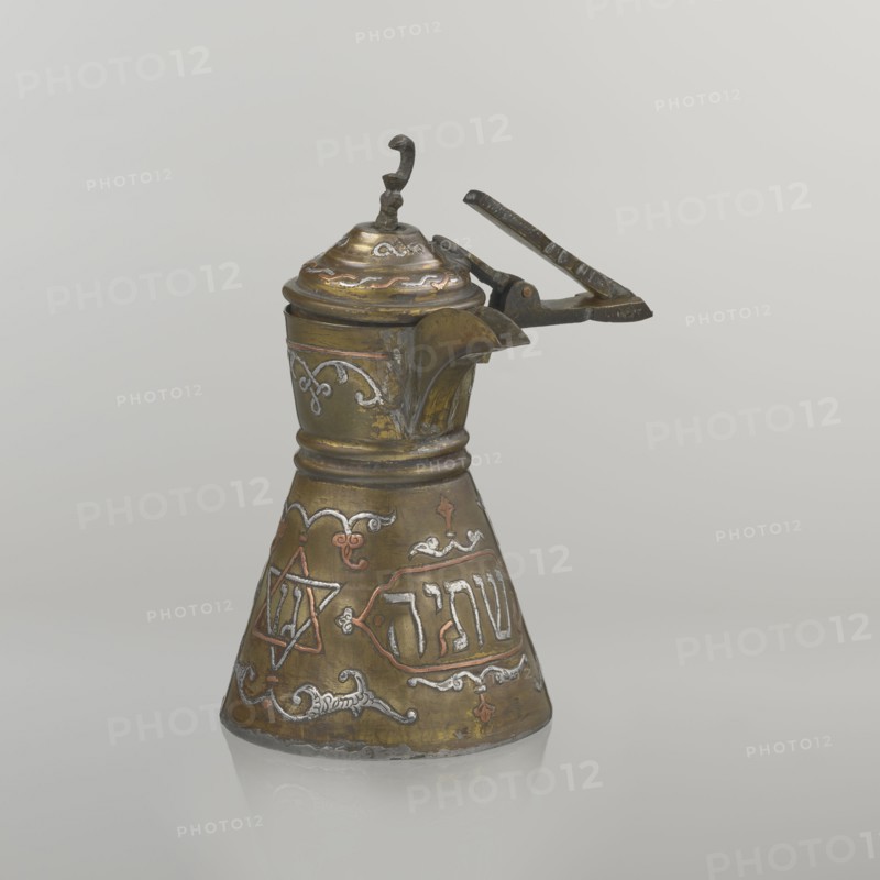 Coffee pot, gift, Arabic and Jewish cultural influences; cezve coffee vessel, beginning of the 20th century, Palestine. Decoration: Hebrew inscriptions inlaid in copper, Star of Solomon motif six-pointed star, arabesque patterns, crescent symbol. Hinged lid hinge halfway down the handle; lid topped with a crescent. Inscriptions deciphered: words including drinking; in the hexagonal star Gan Eden, garden; in the cartouche for celebration; under the handle, in the Seal of Solomon/Star of David Mim from the word water. Vessel: height 15.5 cm, width 15.5 cm, diameter 8.7 cm., A metal pitcher with a hinged lid and a curved spout sits against a plain light gray background; the pitcher has a flared conical base and a narrower neck with decorative raised bands, a small ornate finial on the lid, and a simple lever handle attached near the top. The surface shows a mix of metallic colors including brassy gold, darker bronze, and accents of silver and copper, with areas of darker patina and wear. ...