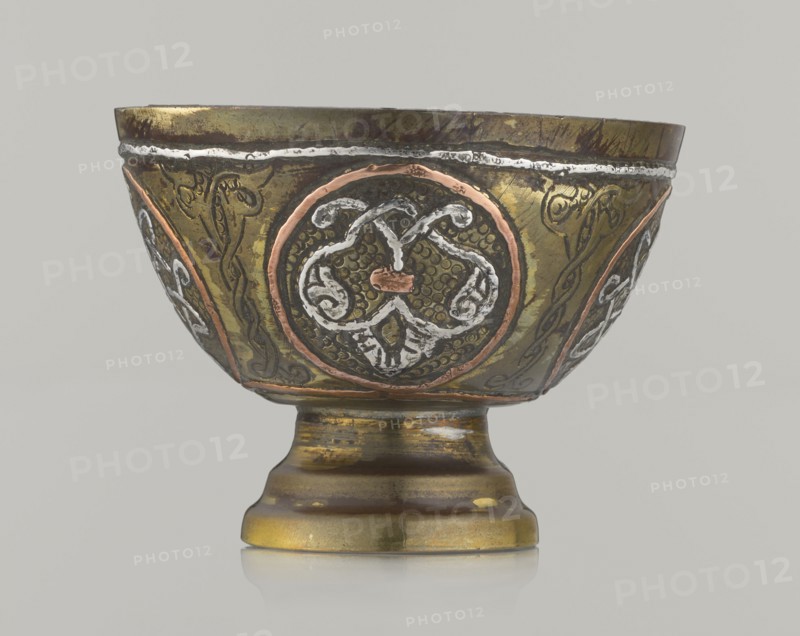 Bowl;, 20th century; Middle East, possibly Palestine; Islamic/Arabic; vessel, height 4.3 cm, diameter 5.8 cm; decorated with braided and arabesque motifs in cartouches on a punched background., A metallic bowl on a pedestal with decorative circular medallions and ornate patterns around its exterior, showing a mix of metallic gold, brass, copper, silver, and dark brown tones with areas of wear and patina; the bowl has raised and engraved motifs in contrasting metals, a flared rim with a thin silver-colored band near the top, and a rounded foot with horizontal stepped profiles, all set against a plain light gray background.