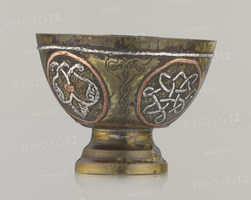 Bowl, Islamic art, Arabic culture; 20th century, possibly Palestine; vessel, height 4.3 cm, diameter 5.8 cm; decoration with braided and arabesque motifs in cartouches, background punched., A metal goblet sits against a plain light gray background, showing a rounded bowl on a short flared stem and circular base. The surface of the goblet displays a metallic olive-green patina with areas of darker brown and worn gold tones, and there are copper-colored rings around two oval medallions on the bowl. Each medallion contains raised decorative knotwork rendered in a bright silvery-white metal, contrasted against a textured dark background. A thin silvery band runs near the rim, and faint engraved lines and darker linear markings appear across the body. The base shows a mix of gold, brown, and greenish hues with patches of darker wear and a small area of exposed lighter metal. Overall the palette includes green, brown, gold, copper, silver, white, and dark gray.