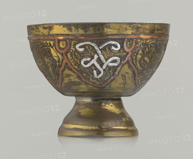 Bowl, gift, Islamic art, Arabic culture, arabesque ornament, geometric motifs, punched background, 20th century, Middle East, possibly Palestine, vessel; height 4.3 cm, diameter 5.6 cm., A decorative chalice-like bowl sits on a rounded foot, showing a metallic surface with worn areas and a patina. The bowl features embossed and engraved patterns around its exterior, including curving, interlaced motifs highlighted in a lighter metallic tone and bordered with a darker outline. There are raised linear decorations in a reddish-copper color tracing triangular panels around the body, and the background of the panels has textured dotwork and engraved designs in darker shades. The overall colors visible are gold, bronze, copper, dark brown, and a lighter silvery-white accent on the central motif, set against a pale neutral background.