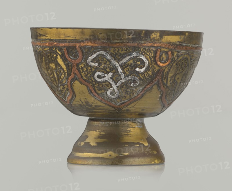 Bowl, 20th century, Middle East possibly Palestine. Ceramic vessel with arabesque and geometric motifs and punched background; height 4.3 cm, diameter 5.6 cm., A decorative metal bowl on a pedestal with an aged, reflective surface showing a mix of gold, brass, copper, and silver tones; the bowl exterior features raised, curving ornamental patterns highlighted in copper and silver against a darker gold background, with textured dotted areas and stylized motifs around the rim; the pedestal base shows worn patches of gold and darker bronze, with irregular areas of patina and surface wear; the overall color palette includes gold, brass, copper, silver, bronze, and muted gray, set against a pale neutral backdrop.