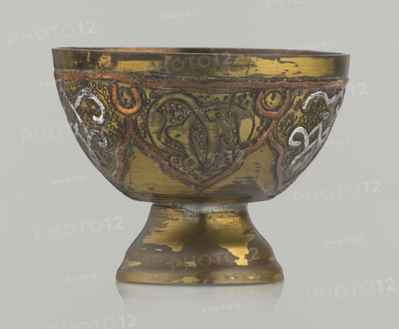 Bowl, Islamic art; Arabic; 20th century, possibly Palestine; vessel with arabesque and geometric motifs, punched background; height 4.3 cm, diameter 5.6 cm., A metal bowl on a short pedestal base with decorative raised patterns and inlaid accents, showing worn and scratched surfaces. The bowl and base are primarily gold and brass tones with patches of darker brownish areas where the finish is worn. There are raised decorative lines and motifs in coppery orange and silvery white against the gold background, with some darker engraved details creating texture. The interior rim and top edge show darker brass coloration, and the overall finish has an aged, patinated look with irregular areas of shine and dullness. The background behind the object is a neutral light gray.