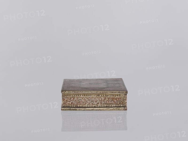Box with lid. Decorated on the sides with plant twigs. Paste colors: red, orange, black paste pressed into engraved patterns. 1st half of the 20th century. North India. Indian art; smoking accessories; casket/box. Materials/technique: applied art, plant and geometric ornament. Dimensions: height 2.8 cm, width 12.8 cm, depth 9.0 cm., A small rectangular box sits centered on a smooth reflective surface, showing a faint mirrored reflection beneath it; the box has an ornate patterned band along its front edge with etched decorative motifs and a slightly raised border, and the lid appears flat and slightly weathered; visible colors include muted gold, brown, reddish-brown, and hints of gray against a pale light-gray background.