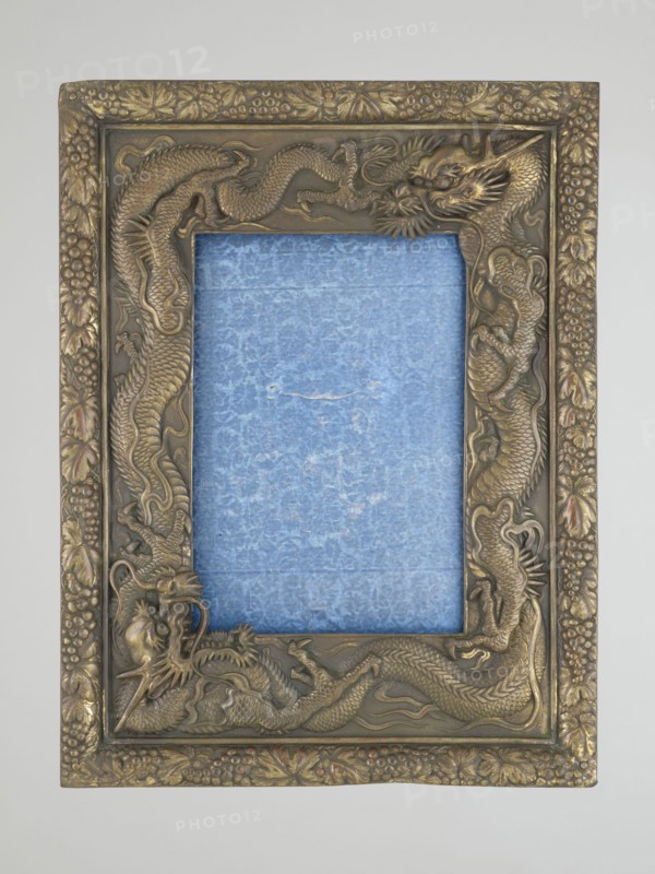 Frame with glass and raised back support covered with blue silk with floral patterns; originally for displaying. Frame decoration: relief patterns of dragons and grape clusters. 20th century, China. Materials: frame with glass and raised back support covered with silk. Height 19.5 cm; width 12.0 cm; depth 1.7 cm., A rectangular ornate picture frame with a raised dragon motif surrounding a textured inner panel, the frame showing metallic tones of gold and bronze with some darker brown shading and subtle hints of reddish highlights; the inner panel is filled with a textured blue material that has lighter and darker blue variations and a small worn area, and the outer border of the frame features carved leaf and berry decorations in matching gold and bronze hues.