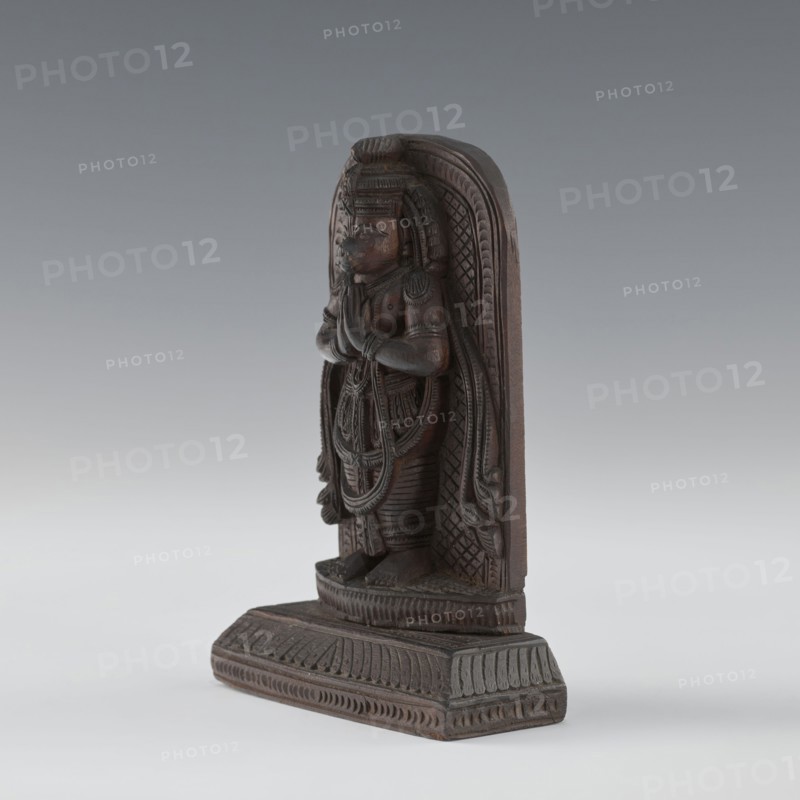 Hanuman, Indian art; South Asian/Indian culture; subject: monkeys; Vaishnavism; Rama; Sita; Ramayana; Hinduism; depiction includes armlets and servants; 1st half of the 20th century, India; bas-relief; height 9.8 cm, width 8.8 cm, A carved wooden statuette stands on a rectangular base, showing a figure in a frontal pose with hands together at chest level; the carving has detailed patterns on the figure’s clothing and headdress, layered necklaces and arm ornaments, and carved textures forming a patterned arch behind the figure. The object displays a range of brown tones in the wood, with some darker brown in recessed areas and lighter brown on raised surfaces, set against a smooth gradient background of gray and pale gray.