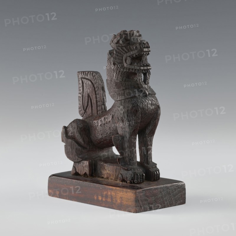 Lion;, figurine; Indian/South Asian culture; animals; claws; 1st half of the 20th century; India, / Sri Lanka ; height 13.2 cm; width 9.7 cm, A carved wooden sculpture of a stylized animal with a raised head and open mouth sits on a rectangular wooden base; the figure has a textured surface with visible carving marks, a mane-like neck, pronounced front legs with carved paws, a curled tail, and a vertical back panel rising behind the body. The visible colors are dark brown and lighter brown variations on the wood, with grey background tones.