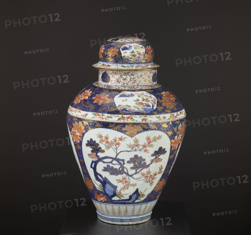 Vase tsubo with cover, Japanese, Imari style, Edo period 1603–1868, decorated with plum trees, bamboos, peonies, pine trees, plant tendrils, meander ornament, and genre scenes; Arita, Japan; 1690–1730; vessel; height 68.5 cm, diameter 45.0 cm., A lidded ceramic vase sits against a dark background, showing a bulbous body narrowing to a short foot and topped with a rounded lid; the vase displays a decorative palette of deep blue, white, orange, gold, green, and touches of red, with large white cartouche-shaped panels featuring a floral branch motif in dark blue and orange, surrounded by deep blue fields filled with stylized orange flowers and gold foliate patterns, a band with small painted scenes around the shoulder, a flared rim with intricate gold and blue ornament, and a foot rim decorated with blue geometric stripes.