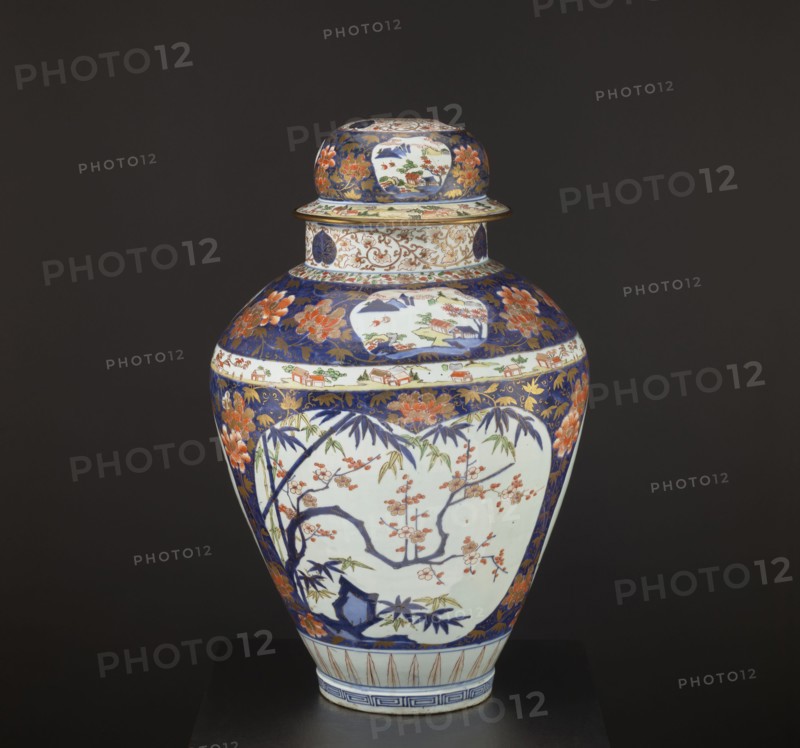 Vase tsubo with cover, Japanese, Imari style, Edo period 1603–1868, circa 1690–1730, Arita, Japan. Porcelain vessel, height 68.5 cm, diameter 45.0 cm. Decorated with floral motifs including plum trees, bamboos, peonies, pine trees, plant tendrils, meander ornament, and genre scenes., A lidded ceramic vase with a rounded body and a narrower neck displayed against a dark background; the vase shows a palette of deep blue, white, orange, green, gold, and touches of brown. The lid and upper shoulder feature deep blue areas decorated with orange floral motifs and gold scrolling, and there are panels of white with painted landscape motifs. The main body has a large white panel bordered by deep blue containing a painted scene of dark blue branches with orange blossoms and green leaves, along with additional floral and bamboo motifs around it. A band of white with small painted house-like shapes encircles the vase near the top, and gold decorative elements appear in the blue areas. The vase sit...