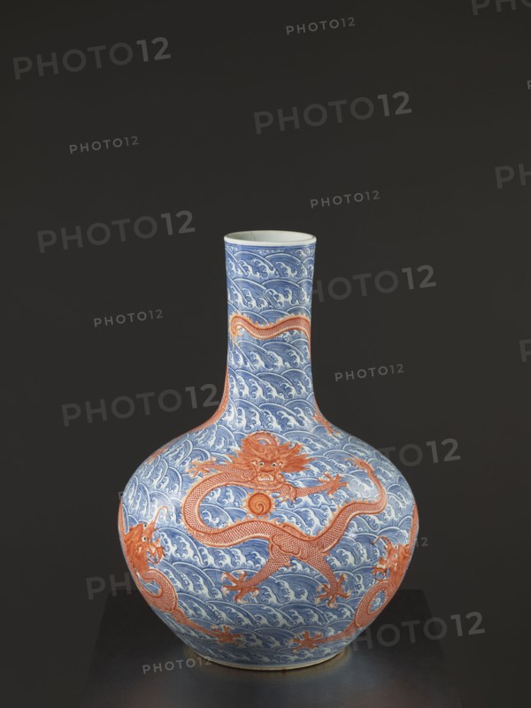 Vase-bottle, Chinese, Qing dynasty Qianlong, 1736–1795, 18th century. Blue-and-white porcelain vessel with bulbous belly, slightly flattened at the top; decoration of dragons with a pearl amid waves/sea waves and pearls. Signed. Height 54 cm; diameter 46 cm., A bulbous ceramic vase with a long narrow neck sits against a dark gray background, decorated with a repeating wave pattern in blue and white and featuring a few large stylized dragons in orange wrapping around the body and neck; the vase rests on a dark surface and shows glossy ceramic highlights and subtle shadowing.