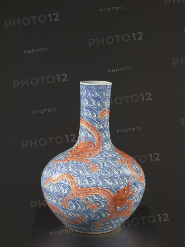 Vase-bottle, Chinese, Qing dynasty, Qianlong period 1736–1795, 18th century. Blue-and-white porcelain; bulbous belly slightly flattened at top; decorated with dragons, dragon with a pearl, waves and sea waves, pearls. Signed. Height 54 cm, diameter 46 cm., A rounded ceramic vase with a long narrow neck sits against a dark background, decorated in blue and white wave patterns covering its surface and accented by a few large orange-red dragon motifs that wrap around the body and neck; the top rim appears pale, and the base shows a touch of warm beige.