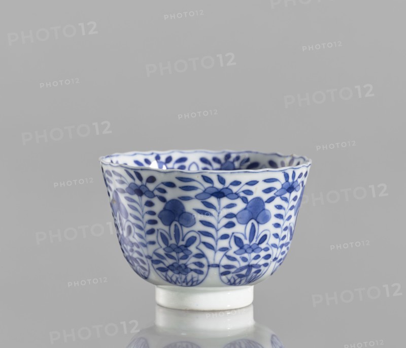 Bowl, Chinese, blue-and-white porcelain, ceramics; floral and plant ornament decoration; conical with serrated rim; Kangxi period 1662–1722, Qing dynasty 1644–1911; China; vessel; height 5.6 cm, diameter 7.9 cm, A small white ceramic cup with blue floral patterns around its exterior sits against a neutral gray background, showing a scalloped rim and a short cylindrical foot; the blue decoration features stylized leaves and flower motifs arranged in repeating vertical bands, and the cup casts a faint reflection on the surface below, with visible colors including white, blue, gray, and a slight off-white on the foot.