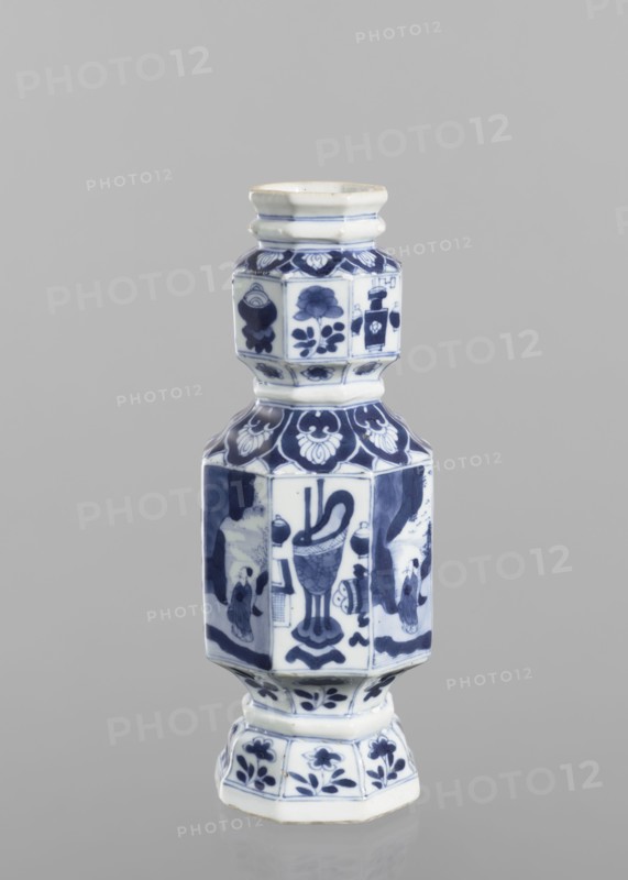 Vase, figurative scenes, ceramics, Chinese art, blue and white porcelain, floral decorations, Qing 1644-1911, decoration with repeating figurative scenes, flower vases and flowers, 18th century, China, vessel, height 13.4 cm, diameter 8.8 cm, A blue-and-white ceramic vessel with a multi-tiered, hexagonal shape sits against a plain gray background; the object displays a glossy white base color with deep blue painted decorative motifs including floral patterns and abstract shapes on each faceted panel, a narrower neck with banded ridges, a wider midsection with stylized figures and ornamental designs, and a flared pedestal base that also features blue floral decorations.