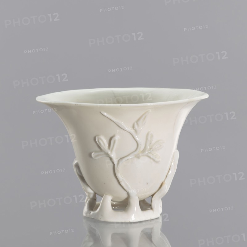 A bowl shaped like rhinoceros-horn carving, Chinese, Qing dynasty 1644–1911, blanc de Chine porcelain with plant ornament and plastic decoration imitating rhinoceros horn, 19th century, Fujian, vessel, height 19 cm, diameter 9.2 cm, A white ceramic cup with a flared rim sits against a pale gray background, showing a smooth glossy surface and a raised branch-and-leaf relief on its side; the cup narrows toward a decorative openwork foot formed by a group of curved supports, and subtle shadows and reflections appear on the surface beneath it.