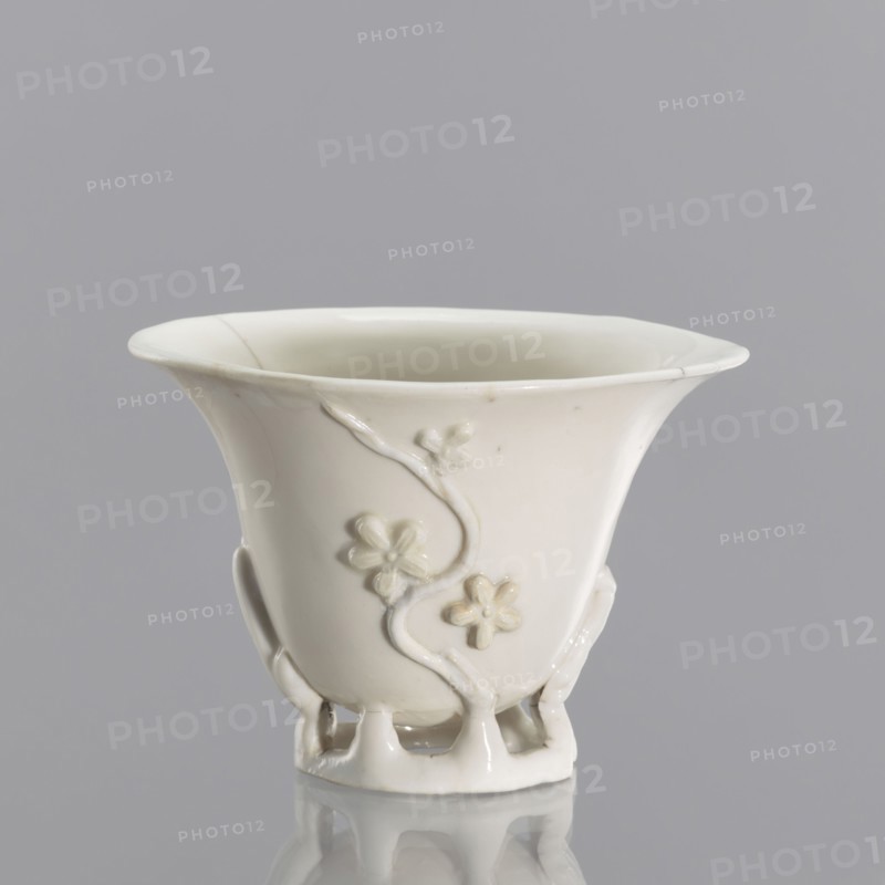 A bowl in a shape reminiscent of a bowl carved in rhinoceros horn; creator, Chinese; Qing dynasty 1644–1911; 19th century; blanc de Chine porcelain with plant ornament and plastic decoration imitating rhinoceros horn carving; from Fujian, China; vessel; height 19 cm, diameter 9.2 cm., A white ceramic cup with a flared rim and a glossy finish sits against a gray background, showing a decorative raised floral vine motif on its exterior and an open lattice-like foot, with subtle shadows and reflections visible on the surface.