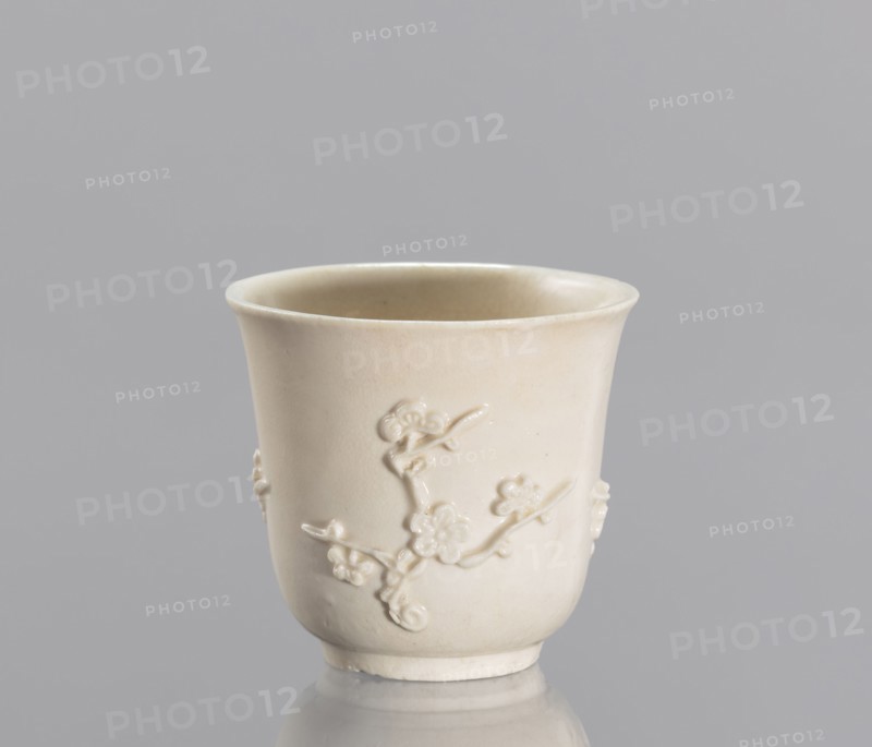 Bowl, plum trees, porcelain blanc de Chine, Chinese art, Chinese culture, decorative vessel with applied/plastic decoration, 19th century China, vessel, height 7 cm, diameter 7.2 cm, A single pale cream-colored ceramic cup sits against a gray background, showing a glossy finish and a slightly flared rim; the cup's surface features a raised floral branch decoration wrapping around its exterior, with the relief petals and stems rendered in the same cream tone, and the cup casts a subtle reflection on the smooth surface beneath it.