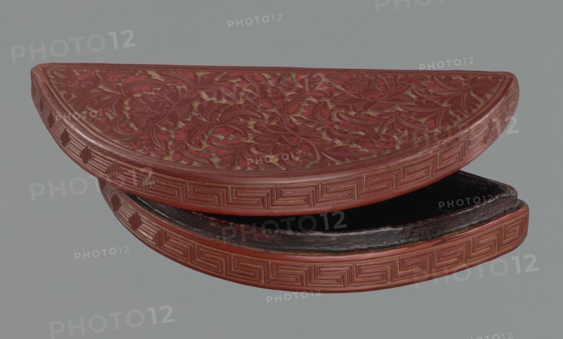 Box with a lid, Chinese art, Chinese culture, Ming 1368–1644, boxes, peonies, meander ornament, leiwen ornament, lemons. Semi-circular box decorated on the lid with a dense pattern composed of peony flowers and leaves. The rim is decorated with a Chinese meander motif. 17th century, China, container. Height 4.4 cm, width 24.0 cm, depth 10.7 cm., A semicircular carved object with intricate floral relief on its top surface and a repeating geometric border along the curved edge, shown partially open to reveal a dark interior. The visible colors include deep red on the carved surfaces, darker reddish-brown along worn areas, black in the interior, and a muted gray background.