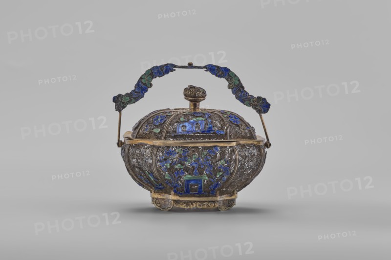 Basket with a lid; maker, Chinese; Canton, China; 19th century; decorated with flowers and ornaments; container; height 9.8 cm, width 10.5 cm, depth 7.5 cm., An ornate metal lidded container with a decorative handle sits against a plain light gray background; the container is bulbous with intricate raised patterns and floral motifs in blue and green enamel set into a tarnished silver or dark metallic body with gold-toned trim and accents, the lid topped by a rounded finial and the hinged handle curved and adorned with blue and green enamel decorations, and the piece rests on a short decorative base with gold-toned elements.