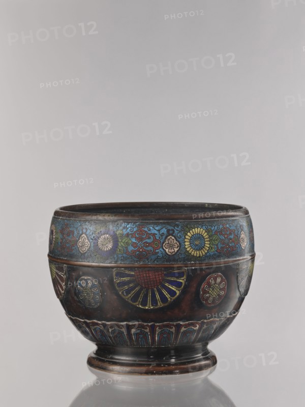 Flowerpot, Shimasa creator/workshop role uncertain, Japanese, Meiji period late 19th century, 1868–1912. Champlevé enamel on bronze jardinière with floral ornament. Vessel; height 30.4 cm, diameter 41.2 cm. Unknown provenance., The image shows a decorative rounded vessel with a short pedestal base, featuring ornate floral and geometric designs in a palette of blue, red, yellow, green, and dark brown, with accents of gold and black; the top band displays a series of stylized flowers on a blue background, the midsection has larger fan-shaped and circular floral motifs with red and blue petals on a dark brown ground, and the lower band contains repeating arch-like patterns in blue and red; the vessel sits on a reflective surface against a neutral pale gray background.