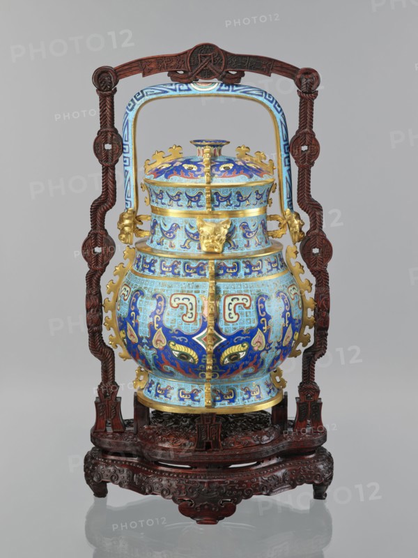 You-type vessel with a lid on a stand;, Chinese, Qing dynasty, Qianlong period 1736–1795. Cloisonné cellular enamel ritual/ceremonial you vessel with elaborate, precise, multi-colored decoration on body and lid depicting a taotie mask and stylized bird images, motifs borrowed from archaic bronzes. Plastic insets on the sides and animal head terminals on the movable handle. Originally used as a vessel for storing alcoholic beverages you, Shang dynasty function. Wooden stand of elongated shape, mattly polished rosewood, decorated with bas-relief symbolic representations bats and spiral ornament. Dimensions: vessel H 39.0 cm, W 24.0 cm, D 19.0 cm; weight 5.6 kg. Stand: H 55.0 cm, W 30.0 cm, D 18.0 cm; weight 1.8 kg., An ornate vessel with a domed lid sits in a carved wooden stand; the vessel is predominantly blue with lighter blue and turquoise sections and gold accents, and it features intricate swirling and geometric enamel patterns in darker blue, red, and touches of green and yellow. ...