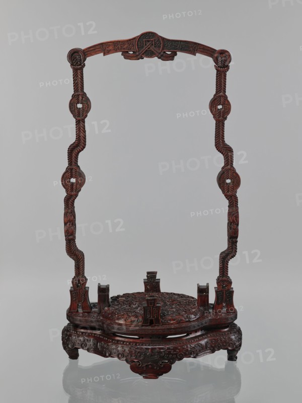 You-type vessel with a lid on a stand. Chinese; Qing dynasty, Qianlong period 1736–1795. Cloisonné cellular enamel ritual/ceremonial you vessel originally used for storing alcoholic beverages form originates in the Shang Dynasty. Elaborate, precise, multi-colored cloisonné decoration on body and lid depicting a taotie mask and stylized bird images, motifs borrowed from archaic bronzes. Plastic insets decorate the sides; animal head terminals flank the movable handle. Stand: mattly polished elongated rosewood base decorated with bas-relief bats and spiral ornament. Dimensions — vessel: H 39.0 cm, W 24.0 cm, D 19.0 cm, weight 5.6 kg; stand: H 55.0 cm, W 30.0 cm, D 18.0 cm, weight 1.8 kg., A carved wooden stand with a dark reddish-brown finish, consisting of an ornate circular base with a raised carved platform and a decorative apron supported by short curved legs, and two tall sinuous vertical posts rising from opposite sides that are joined by a carved arched top; the base and platform ...