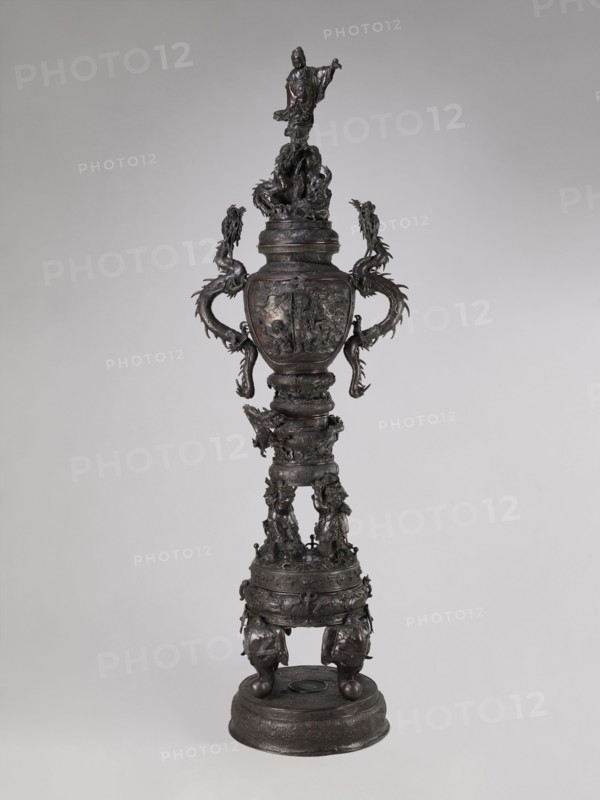 Incense burner koro, Meiji period late 19th century, Japan. Bronze vessel incense burner with dragon-shaped handles; iconography includes dragons, devils, elephants, zodiac signs, peacocks, pine trees, peonies, bodhisattva, and Amaterasu. Height 255.0 cm; width 78.0 cm; depth 46.0 cm., A tall ornate metal urn-like sculpture stands against a plain light-gray background, rendered in dark brown and black tones with subtle lighter highlights; it features a sculpted figure at the top standing on a textured base, flowing drapery, and multiple dragon-like serpentine forms emerging as handles that curve outward and downward; the central body is rounded with detailed relief scenes and decorative bands, leading to an elongated segmented stem adorned with carved figures and ornamental motifs; the lower portion expands into a drum-like base covered in embossed patterns and supported by bulbous feet with animal-head elements, all exhibiting a metallic sheen and intricate surface texture.