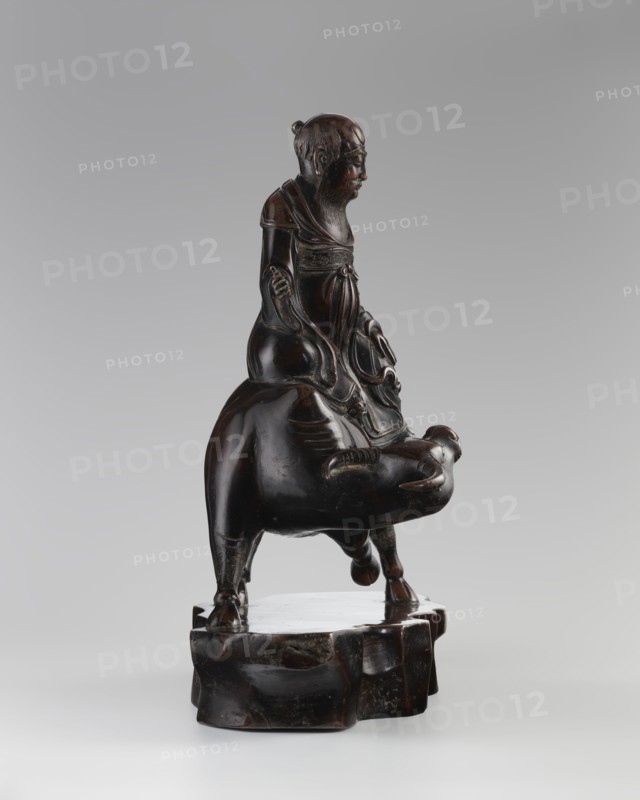 Laozi on a Buffalo, Chinese, Qing dynasty 1644–1911, 19th century. Sculpture/figurine depicting Laozi ca. 550–500 BCE seated on a buffalo; materials not specified. Dimensions: height 39.5 cm, width 41.0 cm, depth 16.5 cm. Subjects: animals, buffaloes., A dark brown sculptural figure made of a glossy material sits on a rounded irregular base of the same color, showing a seated humanlike figure with a long beard and hair tied back, wearing layered flowing garments with carved folds and a tied sash, positioned atop an animal with a rounded body and short legs; the surface displays highlights and subtle variations of darker and lighter brown tones against a neutral gray background.