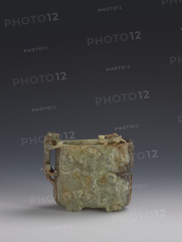 Vessel., Chinese; Qing dynasty 1644–1911. Early 18th century. Gray-green jade, hollowed; bas-relief motifs of clouds and mythological creatures covering the entire surface. Tall vessel on four feet with rounded edges; dragon-shaped handle on the narrower side. Height 9.0 cm; width 14.0 cm; depth 6.5 cm. Icons: dragons, clouds., A small carved object with a square form sits centered against a gradient background that transitions from dark gray at the top to light gray at the bottom; the object is predominantly pale green with areas of brown and darker green, and it features intricate raised swirling patterns and carved details on its surfaces, including a handle on one side with looping shapes and carved decorative elements along the top and corners, and subtle shadows beneath it on the light gray surface.