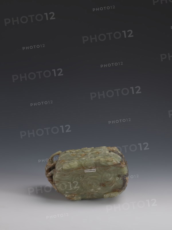 Vessel., Chinese, Qing dynasty 1644–1911, early 18th century. Gray-green jade, hollowed. Tall vessel on four feet with rounded edges; dragon-shaped handle on the narrower side. Entire surface covered in bas-relief motifs of clouds and mythological creatures dragons, clouds. Height 9.0 cm; width 14.0 cm; depth 6.5 cm., A carved object rests on a smooth surface against a dark gray to black gradient background, the object appearing predominantly pale green with areas of brown and darker green mottling; it has a rounded rectangular form with carved swirling patterns across its visible faces and a sculpted handle or loop at one side, and it casts a soft shadow on the surface beneath.