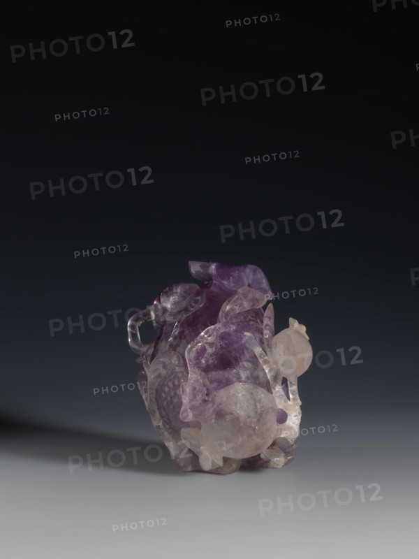 Pomegranate-shaped vessel, Chinese, Qing dynasty 1644–1911, 18th–19th century. Portrays pomegranates and frogs. Vessel; height 10.2 cm, width 9.6 cm, diameter 7.2 cm., A translucent carved object sits on a smooth surface against a dark gradient background, showing a mix of purple, lavender, pale pink, clear, and white tones; the carving has rounded and curved forms with glossy, reflective surfaces and areas of internal cloudiness, with a few bulbous protrusions and looping, ribbon-like elements, as well as textured, scalloped, and faceted details that catch light and cast a soft shadow to one side.