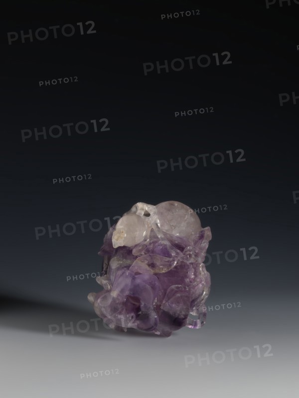 Pomegranate-shaped vessel, Qing dynasty 1644–1911, 18th–19th century, China; porcelain vessel decorated with pomegranates and frogs; height 10.2 cm, width 9.6 cm, diameter 7.2 cm., An irregularly shaped translucent object sits on a smooth surface against a dark gradient background, showing a mix of purple and pale lavender hues with areas of clear, glassy transparency and some cloudy inclusions; the form features rounded bulbous sections and flowing, carved-like contours with glossy highlights and subtle shadows, and the surface reflects light creating small bright spots and softer gleams in tones of purple, clear, and pale gray.