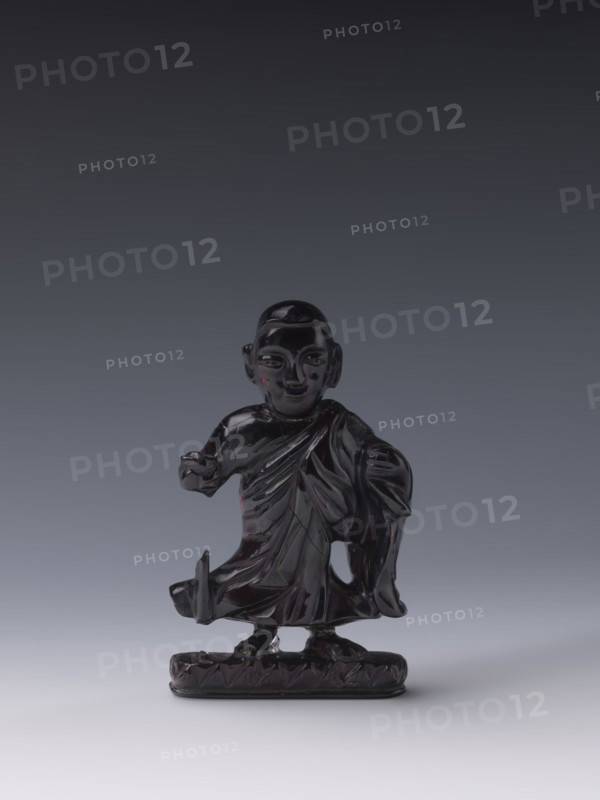 Buddhist monk; unknown creator; Chinese; Qing dynasty 1644–1911, 19th century; Luohan iconography; sculpture, figurine; height 11.8 cm, width 7.1 cm, depth 2.0 cm., A small dark glossy figurine of a standing human figure wearing draped clothing is centered against a smooth gradient background. The figure has a rounded head with defined facial features and ears, and an arm extended slightly forward while the other arm rests by the side. The drapery of the clothing shows carved folds and layered textures, and the figure stands on an oval base with a patterned edge. Visible colors include deep black or very dark brown on the figurine, with subtle highlights of red reflections, and a background gradient of dark gray transitioning to lighter gray toward the bottom.