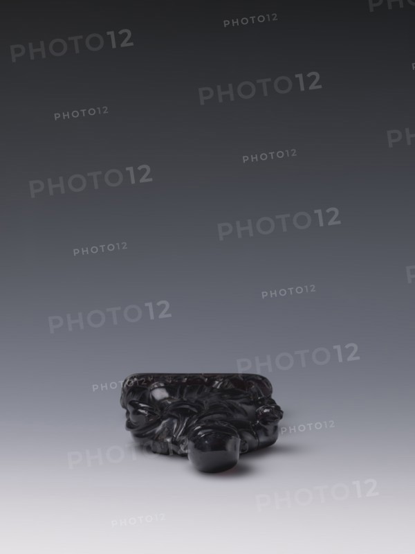 Buddhist monk, Chinese, monks, Qing style, Chinese art, Chinese Buddhism, Qing 1644–1911, Luohan iconography, 19th century, China, sculpture figurine; height 11.8 cm, width 7.1 cm, depth 2.0 cm, A small dark glossy sculpted object sits near the bottom center of the image on a smooth surface, showing a deep black color with highlights and subtle reflections; the object has rounded bulbous forms and flowing carved details suggesting organic shapes, with a slightly irregular silhouette and a polished finish; the background transitions from a lighter gray near the surface to a darker gray toward the top, creating a gradient of gray tones.