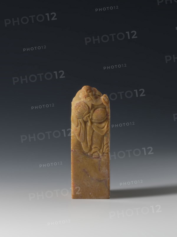 Seal stamp, unknown creator; Chinese; Qing dynasty 1644–1911, 18th–19th century; seal stamp sculpture, Taoist subject depicting the Eight Immortals; inscription on the stamp; materials not specified; dimensions: height 9.3 cm, width 3.2 cm, depth 3.1 cm., A vertical rectangular carved object standing upright on a smooth surface, the material showing warm yellow and brown tones with areas of darker brown and subtle reddish veins; the top portion is sculpted in low relief with a figure wearing flowing robes and holding a fan, the carving exhibiting smooth rounded forms and fine folds, while the lower portion remains mostly plain with natural marbling in brown and reddish hues; the background gradates from a dark charcoal at the top to a lighter gray near the surface, and a soft shadow extends to the right of the object.
