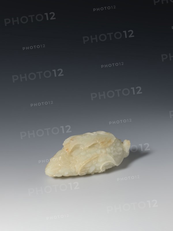 Paperweight - grapevine; maker, pale green jade; sculpture; grapes; writing instrument; Chinese; Qing 1644–1911; 17th–19th century; height 4.0 cm, width 10.3 cm, depth 4.8 cm., A sculpted object made of pale, translucent material with creamy white and light beige tones rests on a smooth surface; the object has an irregular, elongated shape with rounded, overlapping layers and carved, scale-like textures, and it casts a soft shadow on the neutral gray background that gradates from lighter at the bottom to darker toward the top.