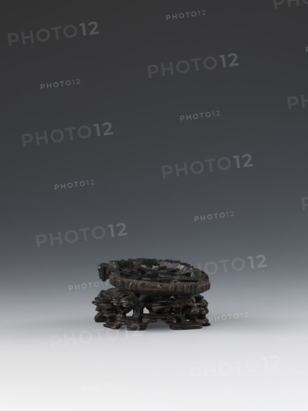 Laozi on base;, Chinese; Qing dynasty 1644–1911; 19th century; taoist subject depicting Laozi ca. 550–500 BCE; sculpture/figurine with decoration on base: rocks, pine trees, flowers, lingzhi mushrooms; materials not specified; dimensions: figurine height 5.2 cm, width 6.3 cm; base height 7.7 cm, width 6.1 cm., A small dark brown carved wooden stand sits near the bottom center of the image, showing a circular top with carved openwork and an intricately carved base with organic, root-like supports; the background above it is a smooth gradient ranging from dark gray at the top to lighter gray toward the bottom, and the surface beneath the stand is a very light gray.