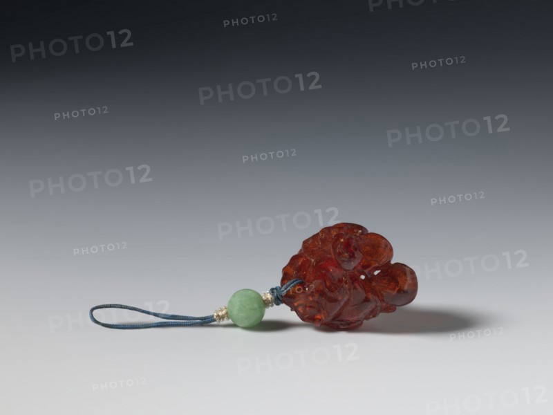 Grape wine pendant, pendant of fossil amber carved with grapes; Chinese, Qing dynasty 1644–1911, 18th century; jewelry; height 3.7 cm, width 3.1 cm., A small carved object made of translucent amber-colored material rests on a smooth surface alongside a short loop of dark blue cord and a round bead of light green stone, with a few small metallic spacer beads near the bead; the background shows a gradient from dark gray at the top to light gray near the bottom.