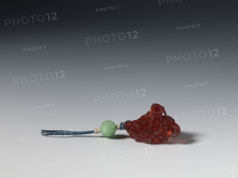 Grape wine pendant, fossil amber; Chinese, Qing dynasty 1644–1911; 18th century; subject: grapes; jewelry pendant; height 3.7 cm, width 3.1 cm; Chinese culture., A small carved pendant lies on a smooth surface, consisting of a translucent reddish-brown carved piece attached to a round green bead and a short loop of blue cord with small pale metallic spacer beads; the pendant casts a soft shadow and the background transitions from light gray at the bottom to dark gray at the top.