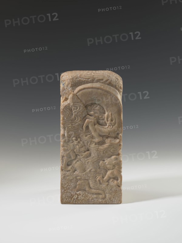 Seal stamp, Chinese art, Chinese culture, Qing dynasty 1644–1911, Qing style, seal stamp, dragon iconography, dragon with a pearl, 18th–19th century, China, sculpture; height 26.5 cm, width 12.0 cm, A rectangular carved stone object stands upright against a neutral gradient background, showing a brownish-beige color with lighter and darker variations; the front surface features shallow relief carvings of sinuous, flowing forms that resemble stylized creatures and cloud-like shapes, with curved arcs and rounded raised areas, and the top is slightly rounded with similar carved motifs and a textured, pitted surface.