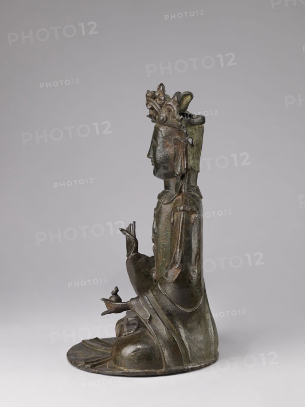 Guanyin, Chinese, 17th century, China, sculpture figurine, bodhisattva and protective deity iconography, Chinese Buddhism; height 43.0 cm, width 23.0 cm., A bronze statue of a seated figure shown in profile against a plain light gray background, with a dark greenish-brown patina and subtle highlights of green; the figure wears a tall ornate headdress with carved decorative elements, has stylized facial features and long earlobes, and wears flowing layered robes with visible folds and rounded drapery; one hand is raised with slender fingers in a gesture while the other hand rests on the lap holding a small rounded object, the figure sits on a rounded base that follows the contours of the robes, and the overall color palette includes dark bronze, green, brown, and muted gray.